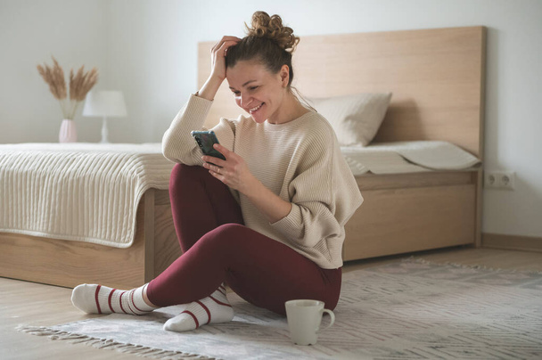 Happy pretty young woman sitting with cellphone on floor in bedroom at cozy home - Fotografie, Obrázek