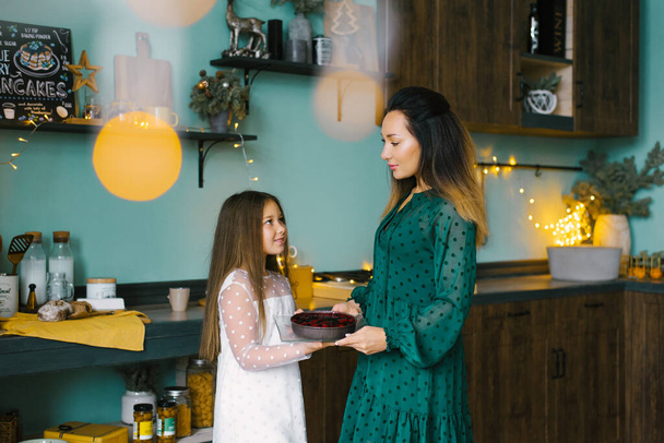 Anne ve kızı mutfakta ellerinde bir Noel pastası tutuyorlar ve birbirlerine gülümsüyorlar. - Fotoğraf, Görsel