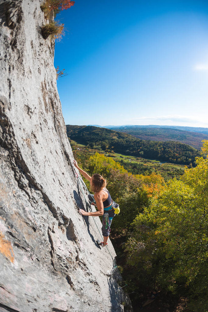 κορίτσι αναρρίχηση βράχου σκαρφαλώνει το βράχο στο φόντο του φθινοπώρου δάσους και του γαλάζιου ουρανού. ένας ορειβάτης περπατά σε μια δύσκολη διαδρομή χρησιμοποιώντας εξοπλισμό. - Φωτογραφία, εικόνα