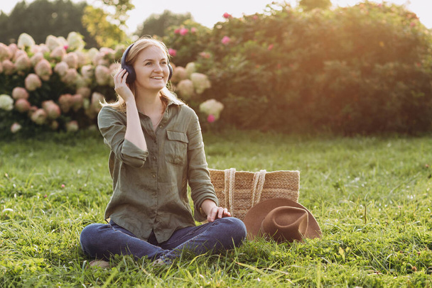 Retrato de una joven rubia feliz escuchando música con auriculares y sonriendo en una hierba verde al aire libre. Amante de la música disfrutando. Conceptos modernos de estilo de vida y relajación. - Foto, Imagen