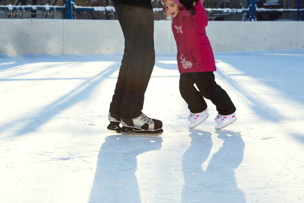 Dad teaches his little daughter to ice skate on a skating rink in the courtyard of multi-storey buildings in the city. Frosty winter sunny day, active winter sports and lifestyle - Фото, изображение