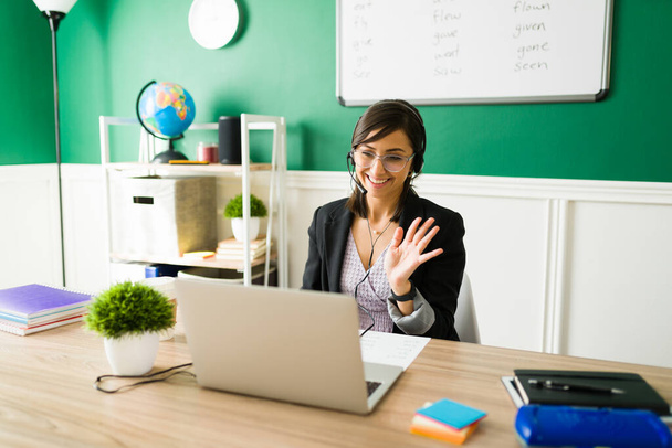Excited female teacher with a headset waving hello to her students and smiling during a virtual class - Photo, Image