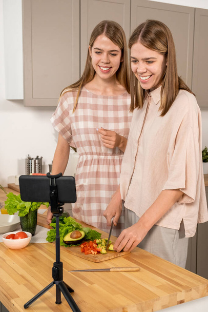 Glückliche süße Teenager-Zwillinge, die vor Smartphone-Kamera Salat kochen - Foto, Bild