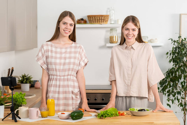 Hermanas gemelas jóvenes felices en ropa casual de pie junto a la mesa de la cocina - Foto, imagen