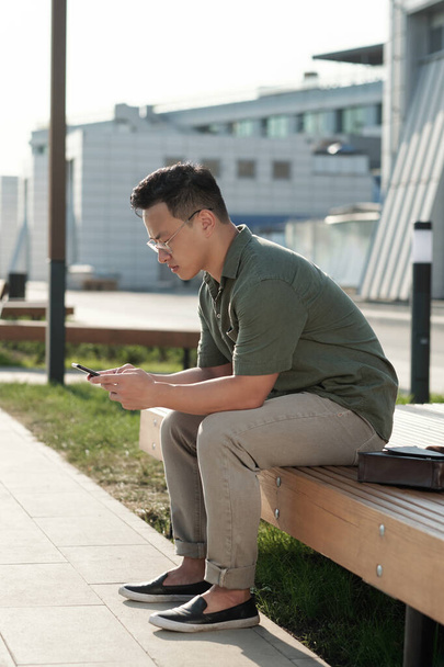 Joven hombre de negocios asiático con teléfono inteligente sentado en el banco - Foto, Imagen
