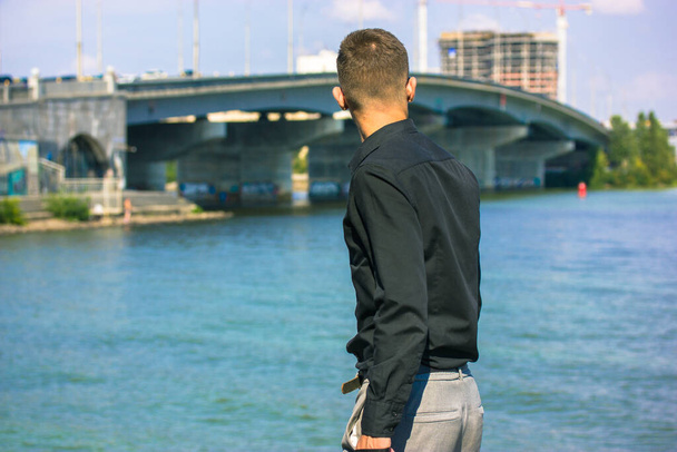 若い男は、黒いシャツの男は、灰色のズボンの堤防の午後には川で、街の風景を探して立っている。橋の後ろの距離に高層マンション建築現場. - 写真・画像