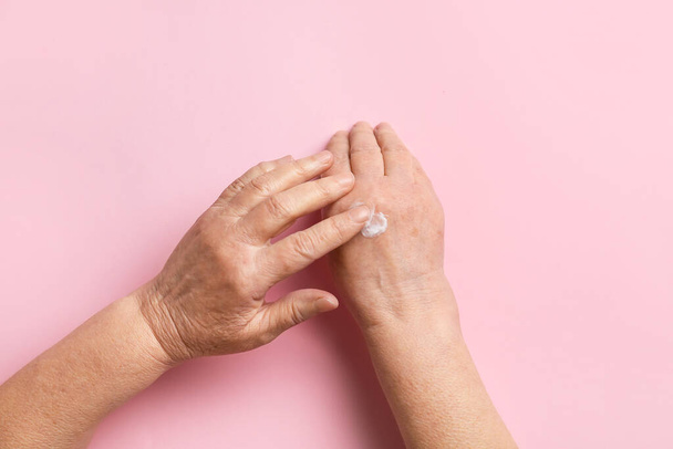 Elderly woman applying cosmetic cream onto her hands against color background - Photo, Image