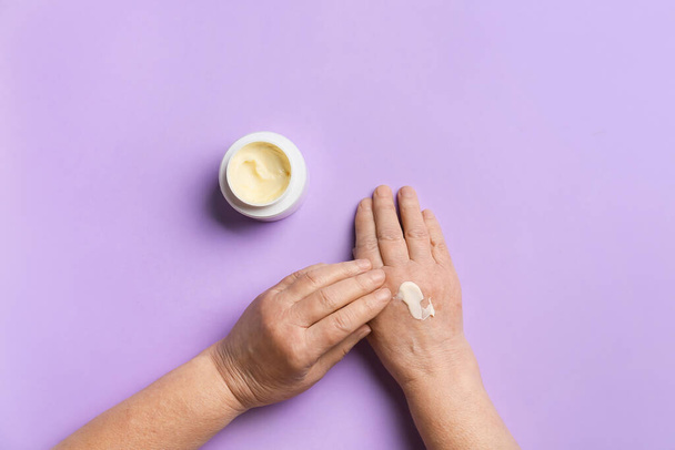 Elderly woman applying cosmetic cream onto her hands against color background - Photo, image