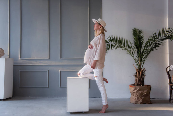 Fashion pregnant young woman in a white hat leaning her leg on a white cube in a gray, stylish studio. The girl in jeans hugs her belly with tenderness. Beautiful, healthy pregnancy. - Photo, image