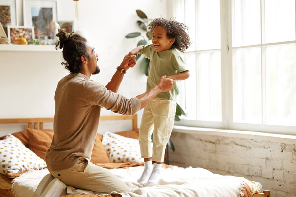 Happy african american father and son jumping on bed and laughing while playing together at home - Φωτογραφία, εικόνα