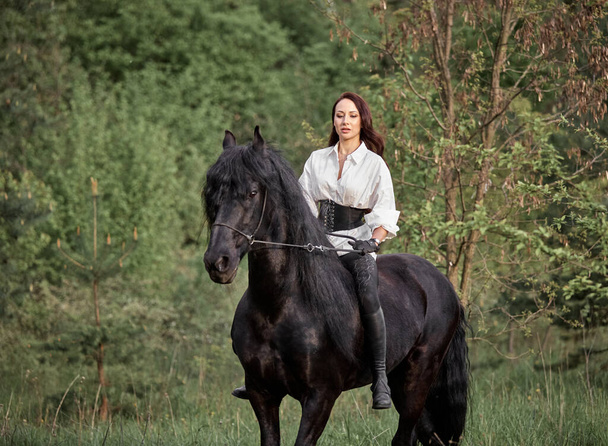 Όμορφο μακρυμάλλικο κορίτσι ιππεύει ένα άλογο Friesian - Φωτογραφία, εικόνα