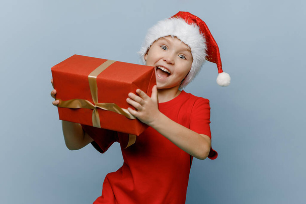 funny white boy of six or seven years old is wearing a Santa hat, holding a gift box in his hands, celebrating the happy New Year 2022. Isolated on a blue background. Merry Christmas gift sale. - Fotó, kép