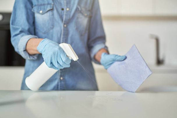 Nahaufnahme weiblicher Hände in Gummihandschuhen mit Waschmittelspray und Tuch beim Reinigen der Küchenoberfläche - Foto, Bild