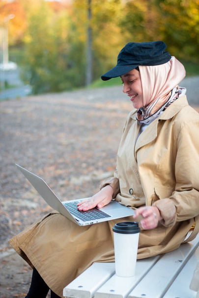 A Muslim woman in a hijab in a beige trench coat sits on a bench with a cup of coffee and a laptop in the park. Muslim business woman. Student. Close plan, face. - Zdjęcie, obraz