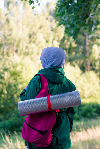 A Muslim woman in a green jacket and a blue hijab with a backpack and a hiking mat in the forest. Hiking in the mountains. - Fotografie, Obrázek