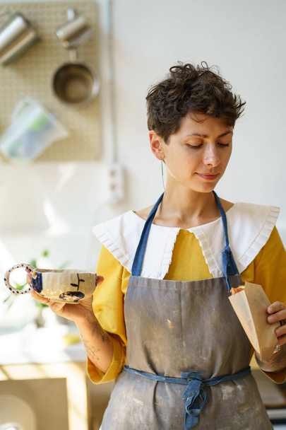 Satisfait jeune femme céramiste boire du café à la main tasse de potier et a collation en atelier de poterie - Photo, image