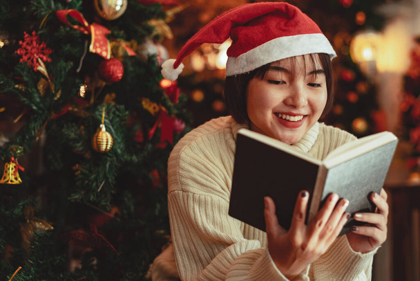 młoda kobieta przed choinką czyta książkę w domu z dekoracją świąteczną - Zdjęcie, obraz