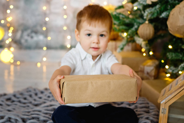 選択的フォーカス。男の子が床に座っていて、ガーランドの照明を背景にクリスマスのために装飾された部屋のギフトボックスがあります。彼はカメラに贈り物を渡す. - 写真・画像