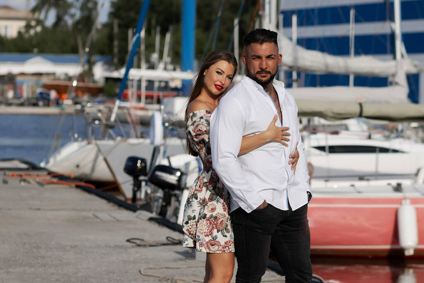 Loving couple in trendy outfits embracing while standing in port with boats in summer - Photo, image