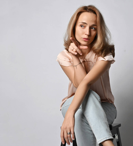 Retrato de una joven rubia en blusa y jeans sentada con las piernas cruzadas señalando con el dedo y mirando a un lado - Foto, imagen