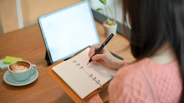 Gros plan Jeune femme tenir un cahier et écrire quelque chose sur le papier tout en regardant l'écran de la tablette. - Photo, image
