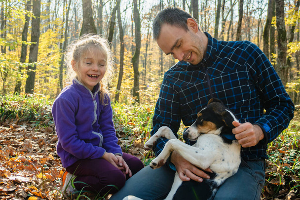 A man in plaid coffee and jeans is stroking a dog lying on his lap. A girl is sitting next to him. People are smiling. Father and daughter in the autumn forest. The concept of a happy family - Valokuva, kuva