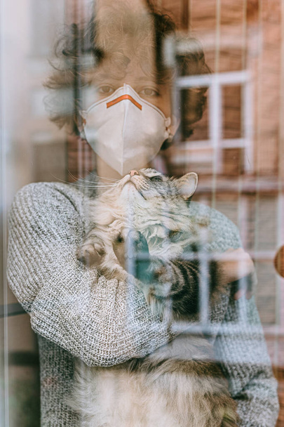 tienermeisje krullend haar trieste bruine ogen beschermende medische masker met grijze pluizige kat haar armen kijkt uit door glazen raam. Quarantaine, zelfisolatie, ziekenhuis. Abstracte refractie licht defocus - Foto, afbeelding