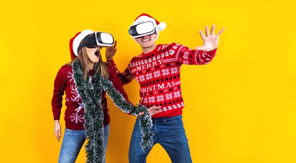 joven pareja latina gamer en realidad virtual aparato de auriculares con sombrero de Santa y suéter de Navidad sobre fondo amarillo. Concepto de tecnología navideña e invernal en México América Latina - Foto, Imagen