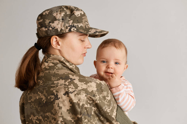 Caucasian military mother wearing camouflage uniform and hat, posing with her infant baby, haven't seen her daughter long time, missing her kid while served in army. - Valokuva, kuva