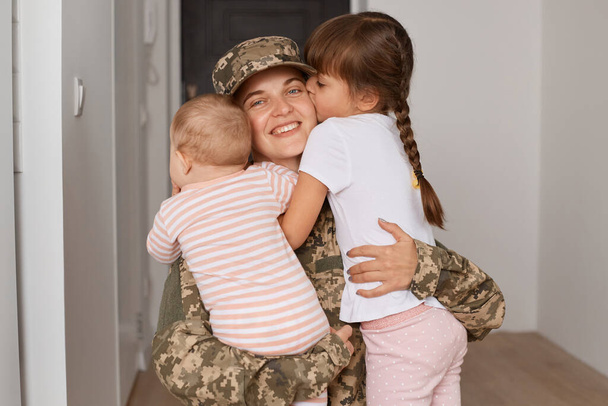 Внутренний снимок патриотичной молодой женщины-солдата в камуфляжной форме, возвращающейся домой после службы в армии и обнимающей своих детей с любовью. - Фото, изображение