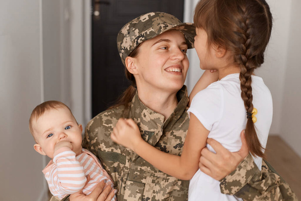 Closeup portrait of happy military mother wearing camouflage uniform and hat, hugging her daughters while returning home after served in army, expressing happiness. - Photo, image