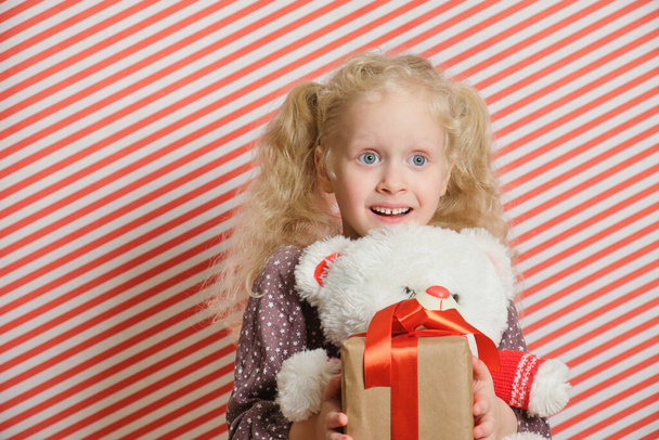 blond preschooler girl with white teddy bear m gift with red ribbon, red striped background, holiday gifts, boxing day, christmas, womens day, birthday party - Φωτογραφία, εικόνα