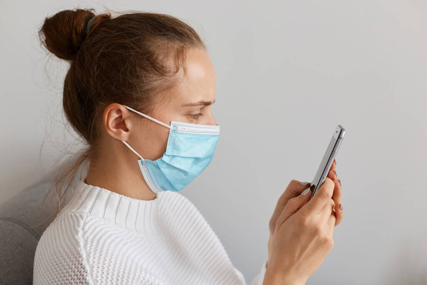 Side view of sad ill woman wearing white sweater and surgical protective mask mask using smart phone, surfing internet or checking social networks while sitting on sofa on quarantine. - Valokuva, kuva