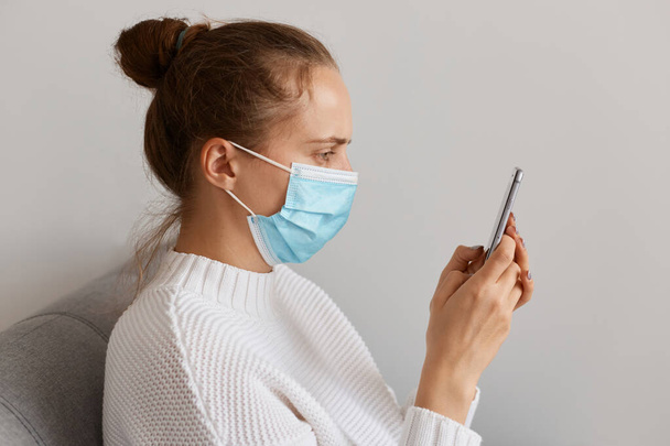 Side view of unhealthy woman wearing white sweater and medical mask, holding cell phone and looking at gadget display, checking email, searching information about treating influenza. - Valokuva, kuva