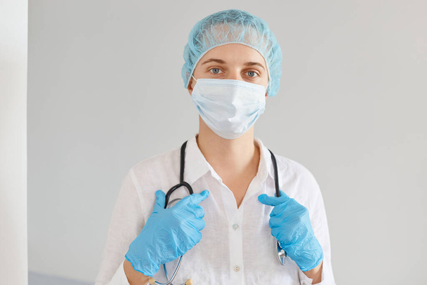 Good looking young adult Caucasian woman doctor wearing medical cap, gloves and protective mask, standing with calm facial expression, waiting her patients. - Foto, immagini