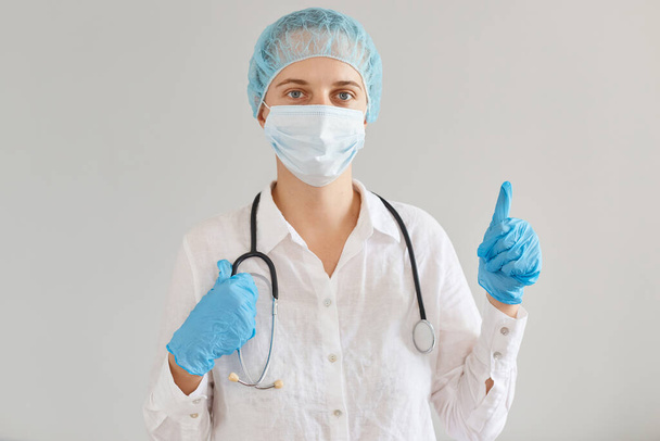 Indoor shot of positive female doctor wearing medical uniform, cap and surgical mask, standing, looking at camera, showing thumb up, approved new methods of virus treatment. - Foto, Imagen