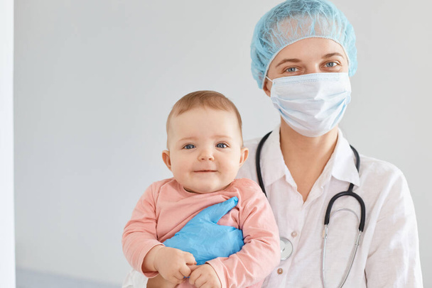 Indoor shot of woman family doctor wearing medical uniform and surgical mask holding infant baby in hands, looking at camera, examines children health. - Φωτογραφία, εικόνα