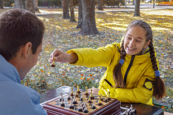 Un ragazzo giovane e una bella ragazza in giacca gialla con le trecce giocano a scacchi all'aperto in un parco autunnale. La ragazza ride e mostra a suo fratello un pezzo di scacchi. Concetto di strategia di successo, vincere il concorso - Foto, immagini