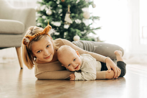 Ein charmantes Mädchen und ihr jüngerer Bruder spielen an Weihnachten oder Silvester gemeinsam auf dem Fußboden neben dem Weihnachtsbaum - Foto, Bild