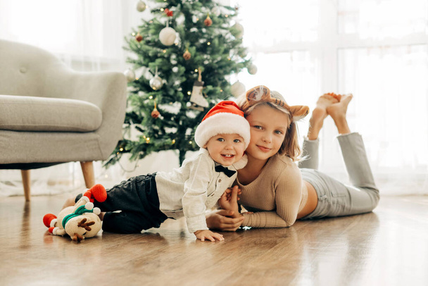 Ritratto di due incantevoli bambini in costume natalizio accanto all'albero di Natale alla vigilia delle vacanze. mood festivo, emozioni positive. Natale e Capodanno - Foto, immagini