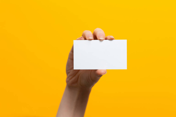Erhobene weibliche Hand mit einer weißen Karte. Attrappe auf. Gelber Hintergrund. Kopierraum. - Foto, Bild