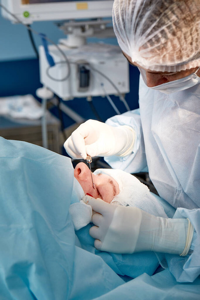 Primo piano del volto di un paziente sottoposto a blefaroplastica. Il chirurgo taglia la palpebra ed esegue manipolazioni con strumenti medici - Foto, immagini