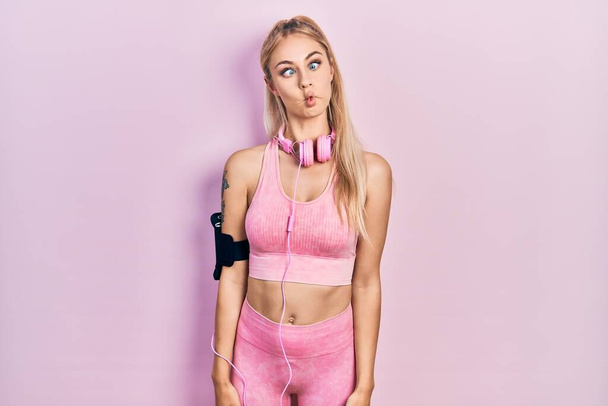 Young beautiful caucasian woman wearing gym clothes and using headphones making fish face with lips, crazy and comical gesture. funny expression.  - Foto, Imagem
