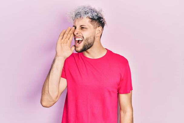 現代的な染めの髪をした若いヒスパニック系の男は、カジュアルなピンクのTシャツを着て大声で叫び、口の上に手で横に叫んでいます。コミュニケーションの概念.  - 写真・画像