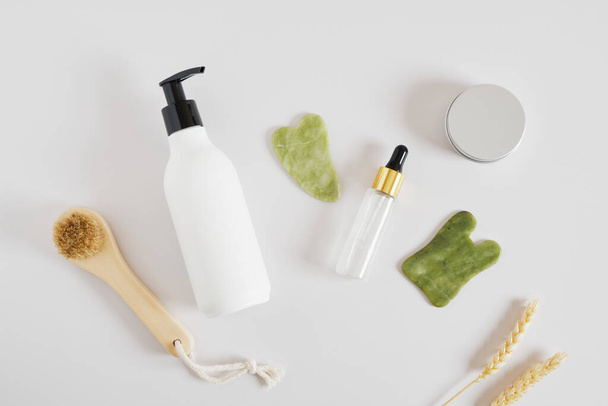 wooden massage brush, aluminum cosmetics jar, cream in a bottle with a dispenser, serum in dropper bottle and a gua sha scraper made of jadeite stone - Fotografie, Obrázek