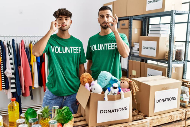 Young gay couple wearing volunteer t shirt at donations stand mouth and lips shut as zip with fingers. secret and silent, taboo talking  - Foto, Bild