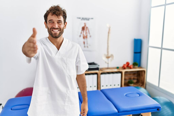 Jonge knappe fysiotherapeut man die werkt bij pijn herstel kliniek glimlachend vriendelijk aanbieden van handdruk als begroeting en gastvrij. succesvol zakendoen.  - Foto, afbeelding