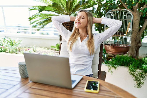 Junge blonde Frau entspannt mit den Händen auf dem Kopf mit Laptop auf der Terrasse. - Foto, Bild