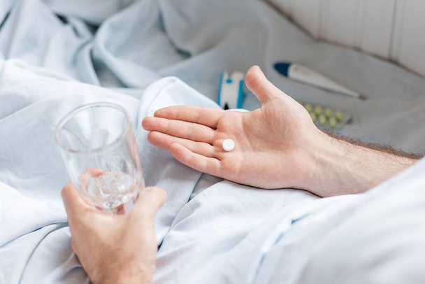 Ein Mann liegt mit einem Glas Wasser im Bett und hält eine Tablette in der Handfläche. Aus nächster Nähe. Ansicht von oben. - Foto, Bild