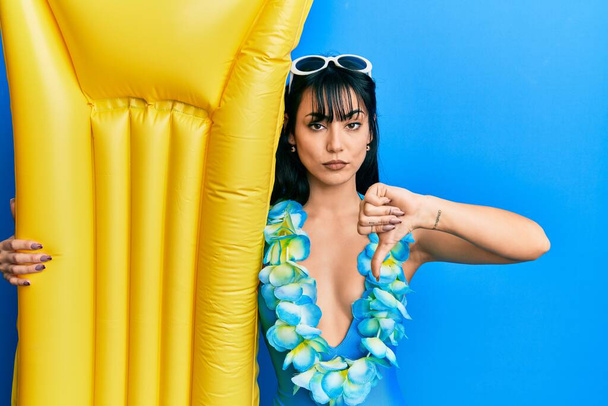 Young brunette woman with bangs wearing swimsuit and holding summer float with angry face, negative sign showing dislike with thumbs down, rejection concept  - Photo, Image